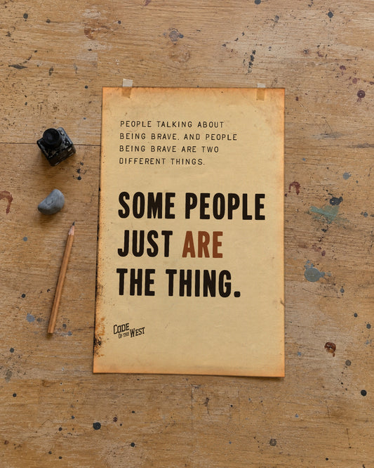 Some People Just Are the Thing poster on aged paper, taped to a paint-flecked wooden drafting table beside a pencil, a bottle of ink and a small stone.