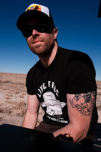 A man gazing off into the distance with a code of the west t shirt on, wearing a black and white trucker hat in front of a blue sky.