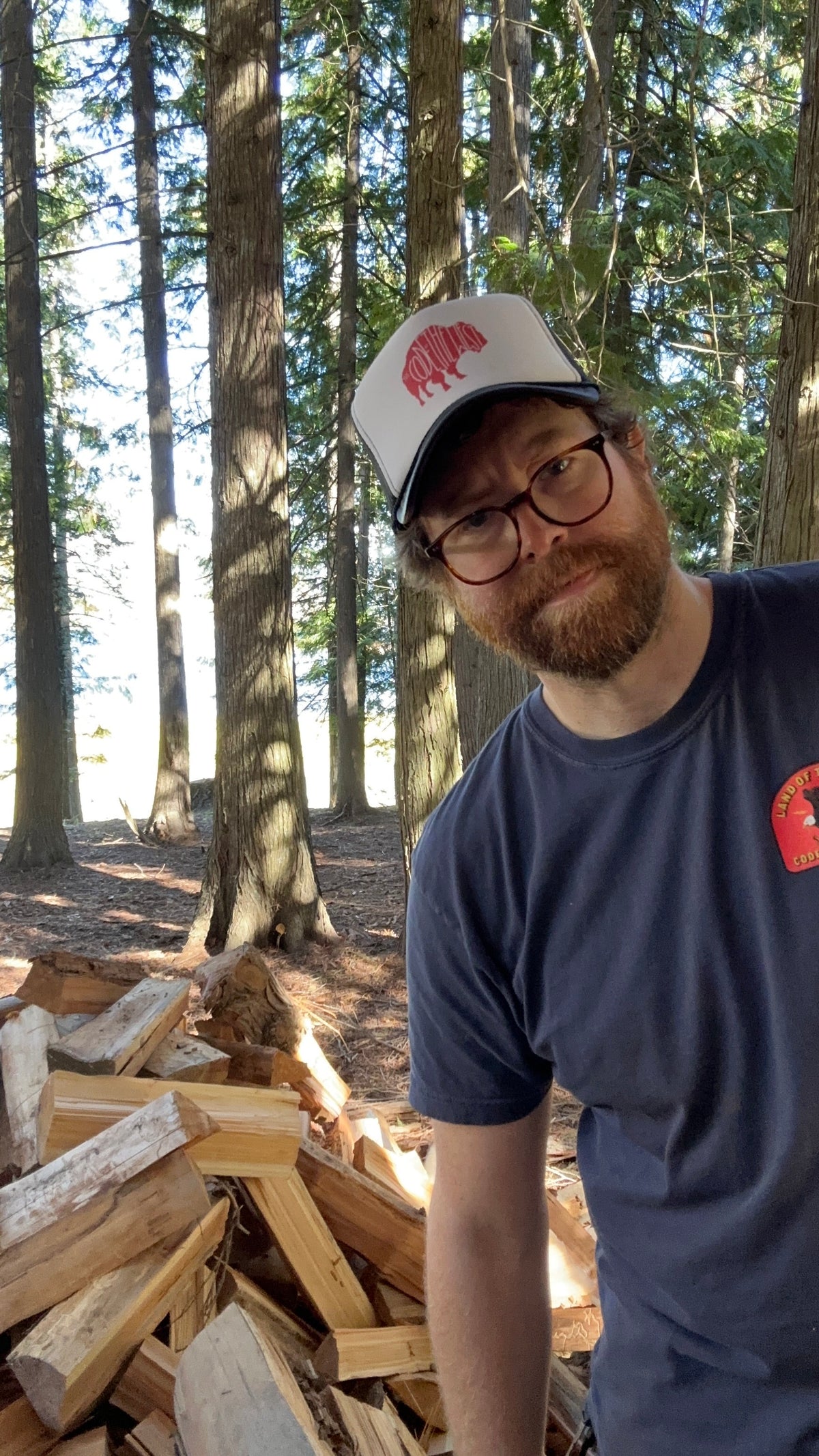 Person wearing the navy Live Free Badge T-shirt beside stacked firewood in the woods