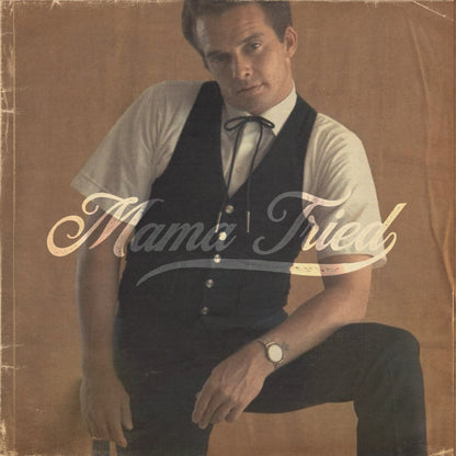 Seated man holding a guitar with cream Mama Tried lettering