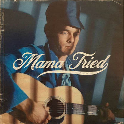 Man wearing a hat and holding a guitar with cream Mama Tried lettering