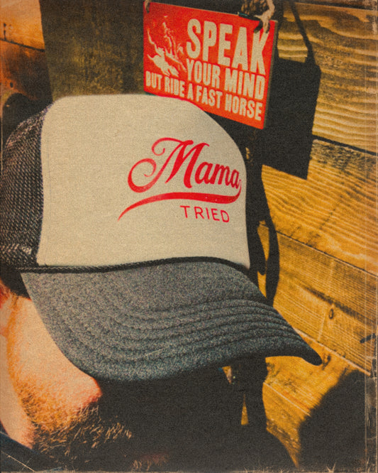 Black-and-white Mama Tried trucker hat with red script, photographed against a wood wall