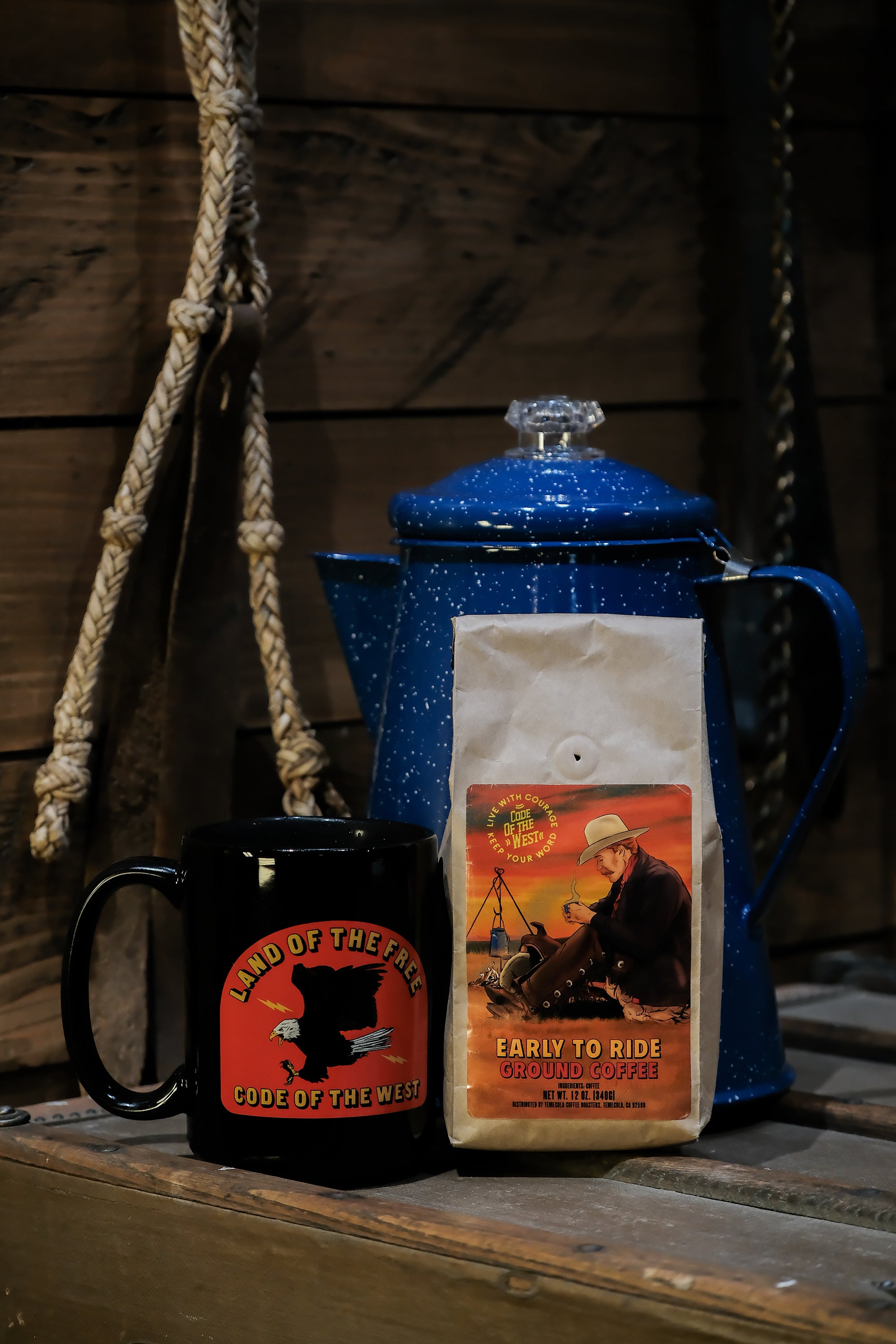 a black mug with a bright red logo showing a bald eagle swooping down with the words Land of The Free and Code of The West, next to an old blue percolator and a bag of code of the west cowboy coffee.